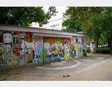 Außenansicht eines Nachbarschaftshauses mit einer Fassade, die vollständig mit bunten Graffiti und Comicillustrationen bedeckt ist. Ein schmaler gepflasterter Weg führt vor dem Gebäude entlang, umgeben von Bäumen und einem kleinen Außenbereich.