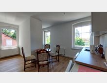 Heller Wohnraum mit einem kleinen runden Holztisch und vier Stühlen, Holzfußboden und zwei großen Fenstern, die Tageslicht und einen Blick auf grüne Bäume ermöglichen; im Vordergrund eine Küchenzeile mit Arbeitsfläche und Küchengeräten.