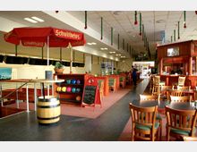 Innenraum einer Bowlinghalle mit Sitzbereichen aus Holz, einer Bar im traditionellen Stil und dekorativen Elementen wie roten Sonnenschirmen und Girlanden. Im Vordergrund befinden sich Bowlingkugeln in einem Regal, während im Hintergrund eine Theke und ein Eingangsbereich sichtbar sind.