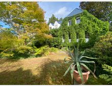 Ein mit Efeu bewachsenes zweistöckiges Haus mit blauen Dachziegeln steht in einem üppigen Garten mit Grasfläche, großen Bäumen und Sträuchern; im Vordergrund befindet sich eine große Agave in einem dekorativen Terrakottatopf.