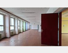 Langer, leerer Flur in einem älteren Gebäude mit großen Fenstern und Heizkörpern entlang der linken Wand sowie abblätternder Farbe an Decke und Wänden. Rechts ist eine geöffnete dunkelrote Tür zu einem angrenzenden Raum zu sehen.