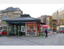 Eingang des U-Bahnhofs Hermannplatz in Berlin mit einem kleinen Gebäude mit grauem Metalldach, das teilweise als Imbissrestaurant "China Box" genutzt wird. Der Platz hat gepflasterten Boden, umliegende historische und moderne Gebäude sowie Fahrradständer und Werbetafeln im Hintergrund.