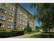 Mehrstöckiges Wohngebäude mit beiger Fassade und weißen Fensterrahmen, umgeben von gepflegtem Grün, Gehweg und Bäumen. Im Hintergrund sind weitere Grünanlagen und Teile eines Parks oder Spielplatzes erkennbar.