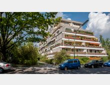 Wohngebäude mit mehreren Stockwerken in moderner Architektur, gekennzeichnet durch große Balkone mit Pflanzen und Markisen. Im Vordergrund stehen geparkte Autos am Straßenrand, während Bäume und Sträucher die Umgebung ergänzen.