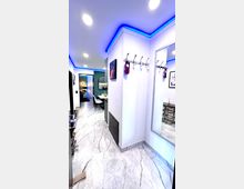 Moderner Flur mit glänzendem, hellgrauem Marmorboden, weiß gestrichenen Wänden und indirekter blauer LED-Deckenbeleuchtung. An der Wand befinden sich Haken, ein großer Spiegel und ein Kunstbild, während der Flur in einen Wohn- oder Essbereich mit dunklen Akzenten führt.