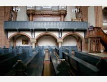Innenraum einer Kirche mit dunklen Holzbänken, roten Backsteinbögen und einer hölzernen Kanzel auf der rechten Seite. Im Hintergrund befindet sich eine Empore mit einer großen Orgel und filigranen Verzierungen.