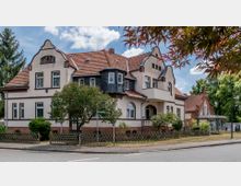 Historisches Wohnhaus mit heller Fassade, roten Dachziegeln und geschwungenen Giebeln, umgeben von Bäumen und einem niedrigen Holzzaun. Neben dem Hauptgebäude steht ein kleineres Gebäude mit ähnlichen architektonischen Merkmalen.
