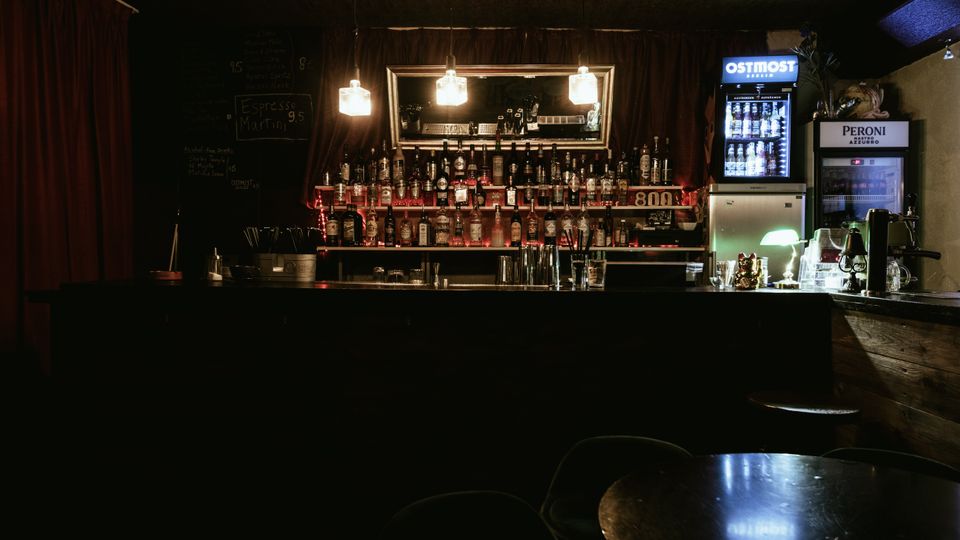 Dimly lit bar interior with a long counter, shelves of liquor bottles, and three hanging pendant lights reflected in a large wall mirror. Red curtains frame the back wall, with a chalkboard menu on the left and illuminated beverage refrigerators on the right.