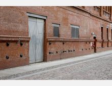 Backsteinfassade eines Gebäudes mit großen grauen Stahltüren, kleinen vergitterten Fenstern und schwarzen Wandbefestigungen. Der Gehweg und die gepflasterte Straße im Vordergrund ergänzen die industrielle Umgebung.