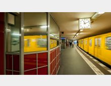 U-Bahn-Station mit gebogenem Bahnsteig, gelber U-Bahn in Bewegung, einer großen analogen Bahnhofsuhr an der Decke und roten Wandverkleidungen aus Glas und Metall. Im Hintergrund sind Personen sichtbar, die auf den Zug warten.