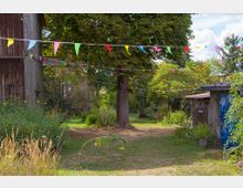Ländlicher Gartenhof mit großem Baum und Wiese, umgeben von üppigem Grün und rustikalen Holzgebäuden. Mehrere bunte Wimpelketten sind quer über den Hof gespannt.