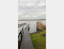 Holzsteg, der über einen ruhigen See führt, umgeben von grüner Wiese und Schilf am Ufer. Im Hintergrund ist eine bewaldete Uferlinie unter einem bewölkten Himmel zu sehen.