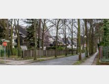 Wohnstraße mit Kopfsteinpflaster, gesäumt von Bäumen ohne Laub. Links moderne Reihenhäuser mit schrägen Dächern, eingefasst durch Zäune und Gärten, rechts ein Gehweg und eine mit Efeu bewachsene Mauer. Straßenschilder und eine Sackgasse werden angezeigt.