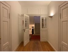 Flur in einem Altbau mit weißen Wänden, Holzdielenboden und Türen im klassischen Stil. Im Hintergrund führt eine Treppe nach oben, flankiert von weißen Doppeltüren mit Glaseinsätzen; rechts eine Wandlampe und ein roter Teppich auf dem Boden.