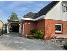 Einfamilienhaus mit roter Klinkerfassade, schwarzem Satteldach und weißem Giebel. Im Vordergrund ein gepflasterter Hof mit Carport, daneben ein kleiner Gartenbereich mit weißer Sitzbank und Tisch.
