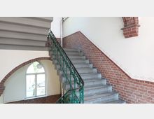 Treppenhaus mit gemauerten Wänden aus rotem Ziegelstein und einem grünen, filigranen Metallgeländer. Im Hintergrund ein großes Rundbogenfenster, das Tageslicht in den Raum lässt, und ein weiß verputzter Wandbereich mit dekorativen Verzierungen.