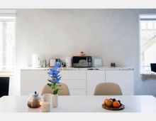 Heller Küchen- und Essbereich mit weißen Unterschränken und marmorierter Arbeitsplatte, auf der kleine Küchengeräte wie Wasserkocher, Toaster und Mikrowelle stehen. Im Vordergrund ein weißer Esstisch mit Teekanne, Vase mit blauen Blumen und einer Obstschale; links und rechts sorgen große Fenster für Tageslicht.