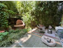 Kleiner Garten mit Bambuspflanzen, einer gepolsterten Liege mit Baldachin, einem modernen Sessel und einem kleinen Beistelltisch. Im Vordergrund liegt ein Hund auf einem ovalen Hundebett auf gepflastertem Boden.