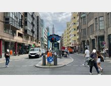 Straßenszene in einer urbanen Umgebung mit mehrstöckigen Wohn- und Geschäftsgebäuden aus verschiedenen Architekturstilen. Im Vordergrund eine Fußgängerinsel mit einer U-Bahn-Eingangstreppe und einer Ampelanlage, umgeben von Autos, Fußgängern und Fahrradfahrern in Bewegung.