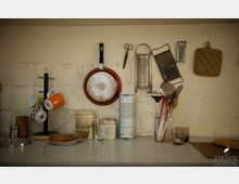 Wand einer einfachen Küche mit Küchenutensilien: Eine rote Pfanne, Reiben, Schere und ein Topflappen hängen an der Wand. Auf der Arbeitsfläche stehen Tassen, Gläser und ein Utensilienhalter mit Kochwerkzeugen. Die Wand ist grob verputzt.