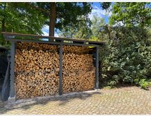 Holzunterstand mit gestapeltem Brennholz in drei Fächern, umgeben von Bäumen und Sträuchern. Der Boden ist mit Pflastersteinen ausgelegt, und im Hintergrund ist ein sonniger, bewaldeter Garten zu sehen.