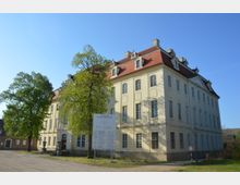 Barockes Schlossgebäude mit heller Fassade, hohen Fenstern und einem roten Mansardendach, umgeben von Bäumen und einer offenen Sandfläche. Im Vordergrund steht ein Schild mit Informationen zum Schloss.