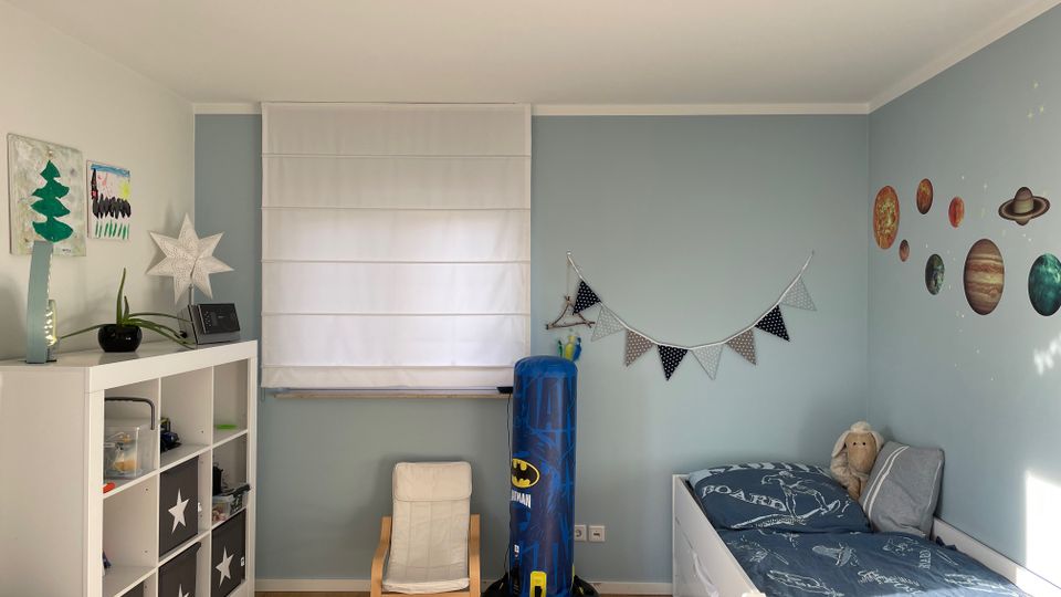 Kinderzimmer mit hellblauen Wänden und einem Holzboden. Links steht ein Regal mit Fächern und dekorativen Aufbewahrungsboxen, daneben ein kleiner weißer Sessel. Rechts ein Kinderbett mit blauer Bettwäsche, an der Wand darüber sind Wandsticker von Planeten angebracht. Ein weißer Rollo bedeckt das Fenster in der Mitte der Wand.