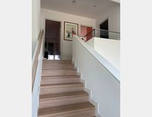 Moderne Innenraumtreppe mit hellen Holzstufen und weißen Wänden, die zu einem oberen Bereich mit Glasgeländer führt. Im Hintergrund befinden sich Türen und gerahmte Bilder an der Wand.