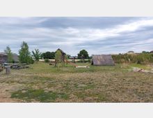 Ländliche Wiese mit einem einfachen Spielplatz, bestehend aus einem hölzernen Kletterhaus mit Rutsche und Schaukeln. Im Hintergrund befinden sich strohgedeckte Hütten sowie vereinzelte Bäume und Sträucher.