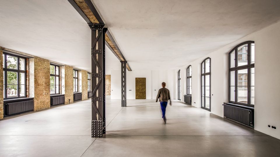 Großer, leerer Loft-Innenraum in einem umgebauten Industriegebäude mit freiliegenden Backsteinwänden, weißen Decken und Stahlstützen. Entlang beider Seiten verlaufen hohe, bogenförmige Fenster mit schwarzen Rahmen und darunterliegenden Heizkörpern über einem glatten Betonboden.