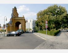 Straßenszene mit einem historischen Backsteinbogenbau auf der linken Seite, daneben geparkte Autos. Rechts ein Gehweg mit großen Bäumen und Gebüsch, mit Verkehrszeichen und einer Kopfsteinpflasterstraße im Vordergrund. Im Hintergrund moderne Gebäude.