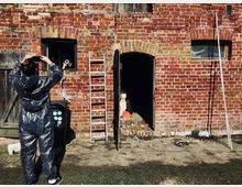 Eine alte Fassade aus roten Ziegelsteinen mit mehreren Bögen, zwei Fenstern, einer offenen Tür und sichtbaren Holz- und Metallgegenständen wie einer Leiter. Im Vordergrund steht eine Person in einem grauen Schutzanzug, die ein Foto macht.