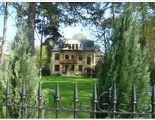 Gelbe Villa mit Mansardendach und großen Fenstern, umgeben von einem gepflegten Garten mit hohen Bäumen und Koniferen. Im Vordergrund steht ein schwarzer schmiedeeiserner Zaun.