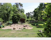 Grüne Gartenlandschaft mit Bäumen im Hintergrund, einem Holzschuppen, Sandhaufen und darauf verstreuten Spielzeugfahrzeugen. Im Vordergrund Kindertraktoren und Bänke auf einer Rasenfläche.