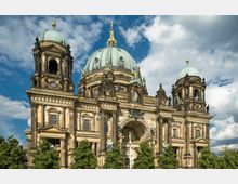 Eine monumentale Kathedrale im neobarocken Stil mit drei grün patinierten Kuppeln und reich verzierten Fassaden. Im Vordergrund sind Bäume und ein wolkiger Himmel im Hintergrund sichtbar.