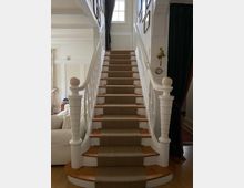 Innenansicht eines Wohnhauses mit einer Holztreppe, die mit einem beigen Teppichläufer belegt ist. Die Treppe hat weiße, verzierte Geländer, dahinter sind Wände mit Bildern und ein Fenster, das natürliches Licht hereinlässt.