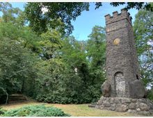 Steinerner Gedenkturm in einem bewaldeten Park, aus dunklen Bruchsteinen mit Zinnen an der Spitze und großen Felsen am Sockel; umgeben von dichtem Laubwerk und einer Rasenfläche.
