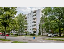 Mehrstöckiges Wohngebäude mit weißer Fassade und Balkonen, umgeben von Bäumen und Grünflächen. Im Vordergrund eine Straße mit geparkten Autos und vereinzelt aufgestellten Straßenschildern.