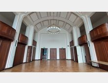 Großer Innenraum mit hohen Decken und einem zentralen Kronleuchter mit kugelförmigen Leuchten, umgeben von Holzvertäfelungen an den Wänden. Drei Holztüren befinden sich an der Stirnwand, flankiert von weißen Säulen und Holzbalkonen.