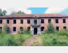 Zweistöckiges, verlassenes Backsteingebäude mit fehlenden Fensterscheiben und Graffiti an der Fassade. Im Vordergrund ist ein überwucherter Bereich mit Gras und Büschen zu sehen.