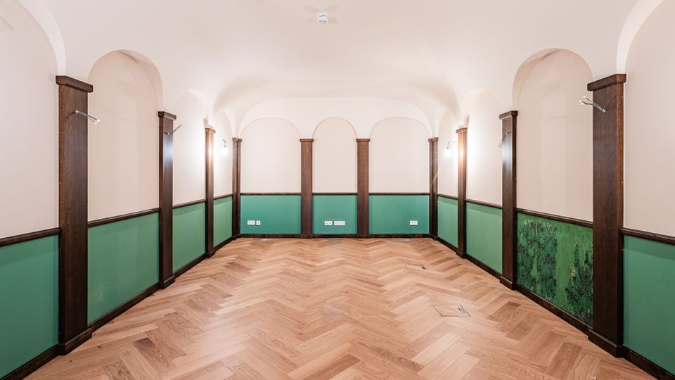 Leerer Innenraum mit weißer Tonnengewölbedecke und rundbogigen Nischen entlang der Wände, gegliedert durch dunkelbraune Holzpfeiler. Unten grüne Wandpaneele und ein heller Fischgrät-Parkettboden, an den Seiten kleine Wandlampen und Steckdosenleisten.