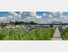 Bootsanleger an einem See mit zahlreichen festgemachten Motorbooten und Segelschiffen, umgeben von Schilf und grüner Vegetation. Im Hintergrund sind eine historische Windmühle, Kirchtürme und Häuser einer Stadt unter einem teils bewölkten Himmel zu sehen.