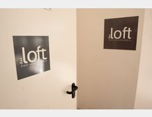 Weiße Innenraumtür und angrenzende Wand mit schwarzen Schildern „das loft – Event Location Berlin“. Die Tür hat eine schwarze Klinke und führt in einen schlichten Flur- oder Eingangsbereich.