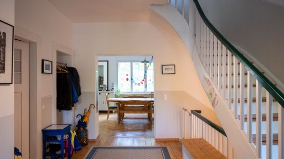 Heller Flur in einem Wohnhaus mit Holzfußboden, Teppichläufer und Garderobenbereich an der linken Wand. Rechts führt eine weiß gestrichene Treppe mit dunkelgrünem Handlauf nach oben, im Hintergrund öffnet sich der Blick in ein Esszimmer mit Holztisch vor einem Fenster.