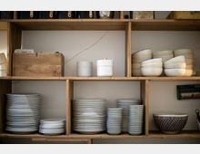 Offenes Holzregal in einer Küche mit gestapeltem weißen Geschirr, darunter Teller, Schüsseln und Tassen. Im Regal befinden sich auch zwei weiße Keramikbehälter, eine dekorative Schale mit Muster und eine Holzkiste.