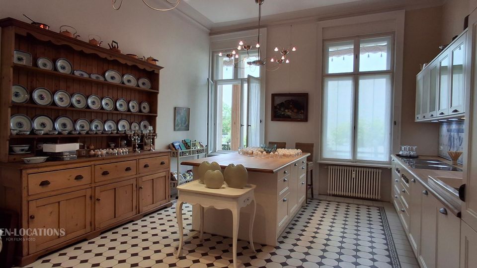 Helle, großzügige Wohnküche mit schwarz-weißem Fliesenboden, zentraler Kücheninsel und hohen Fenstern. Links steht ein großer Holzbuffetschrank mit offenem Tellerregal, rechts eine weiße Einbauküche mit Arbeitsplatte und Oberschränken.