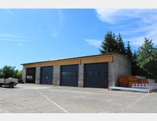 Ein graues Industriegebäude mit drei großen, schwarzen Rolltoren, umgeben von einer betonierten Fläche. Im Vordergrund stehen ein Anhänger, gestapelte Holzplatten und Absperrmaterialien; im Hintergrund sind Bäume sichtbar.