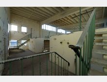 Ein leerer, verfallener Innenraum mit Betonwänden und großen Fenstern, durch die Tageslicht einfällt. Sichtbar sind grüne Metallgeländer entlang mehrerer Treppen, eine Decke mit freiliegenden Trägern und eine beschädigte Wand mit Markierungen.