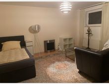 Wohnraum mit neutralen Wänden, einem Einzelbett links, einer Stehlampe, einem hellen Teppich, einem kleinen Couchtisch und einem schwarzen Ledersessel rechts. Ein Fenster mit Vorhängen befindet sich an der Rückwand, daneben ein Heizkörper und ein Pflanzenständer mit Vase.