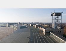 Luftaufnahme eines weitläufigen Gebäudes des ehemaligen Flughafens Tempelhof in Berlin mit seinem geschwungenen Dach und einer großen Freifläche im Vordergrund. Im rechten Teil des Bildes ist ein hoher, offener Metallturm zu sehen, im Hintergrund erstreckt sich die urbane Landschaft Berlins.